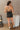 Full body back view of model wearing the Spin Me Around Romper which features washed black denim, two front pockets, a sweetheart neckline, adjustable straps, an open back with corset ties, and a back zipper.
