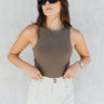 Front view of model wearing the Grace Olive Sleeveless Bodysuit which features dark olive knit fabric, a round neckline, a sleeveless design, and a thong bottom with snap closures.