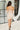 Back view of model wearing the Love Song Beige Off-The-Shoulder Mini Dress that has beige fabric, mini length, a pleated neckline, and off-the-shoulder pleated short sleeves.