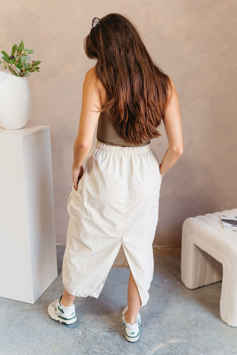 Full body back view of model wearing the Alina Cream Parachute Cargo Midi Skirt that has cream lightweight fabric, midi length, a back slit, two side pockets, an elastic drawstring waistband, and a drawstring hem.