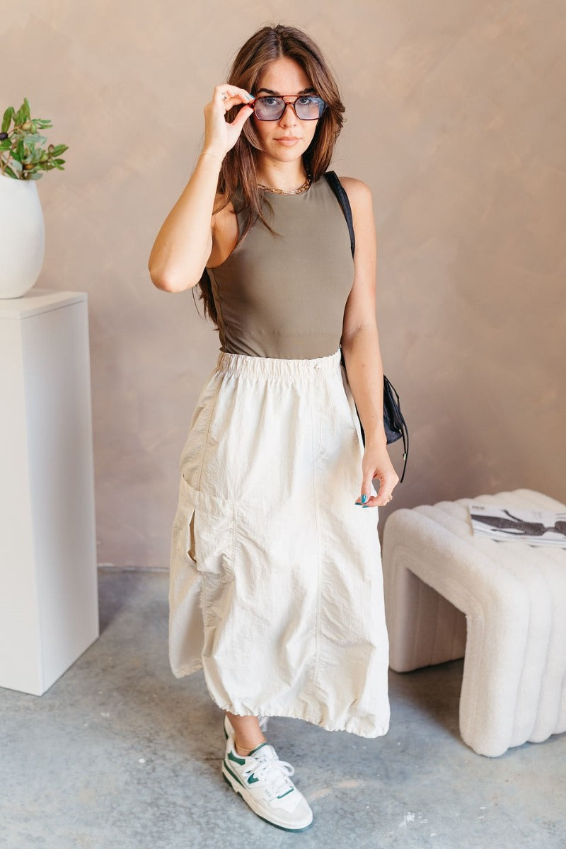 Full body front view of model wearing the Alina Cream Parachute Cargo Midi Skirt that has cream lightweight fabric, midi length, a back slit, two side pockets, an elastic drawstring waistband, and a drawstring hem.