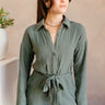 Front view of model wearing the Naomi Dark Green Long Sleeve Romper which features dark green knit fabric, two front pockets, monochromatic button up, collared neckline, tie around the waist and long sleeves with buttoned cuffs.