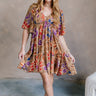 Full body view of model wearing the Harper Multi Floral Mini Dress features light brown fabric with a blue, purple, rust and orange floral pattern, a ruffle hem skirt, a plunge neckline, a tie in the back, and short flare sleeves.