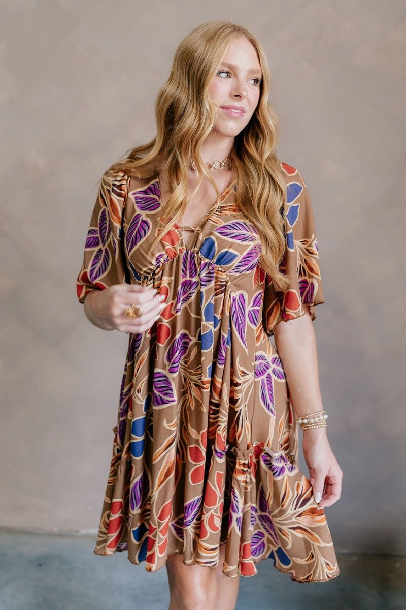 Front view of model wearing the Harper Multi Floral Mini Dress features light brown fabric with a blue, purple, rust and orange floral pattern, a ruffle hem skirt, a plunge neckline, a tie in the back, and short flare sleeves.