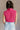 Back view of model wearing the Kelly Pink Cropped Turtleneck Sweater which features hot pink cable knit fabric, a cropped waist, a turtleneck neckline, and a sleeveless design.