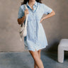 Full body front view of model wearing the Blakeley Distressed Chambray Mini Dress that has light blue chambray fabric, a frayed hem, distressed details, mini length, light tortoise buttons, pockets, a collared neck and short sleeves.