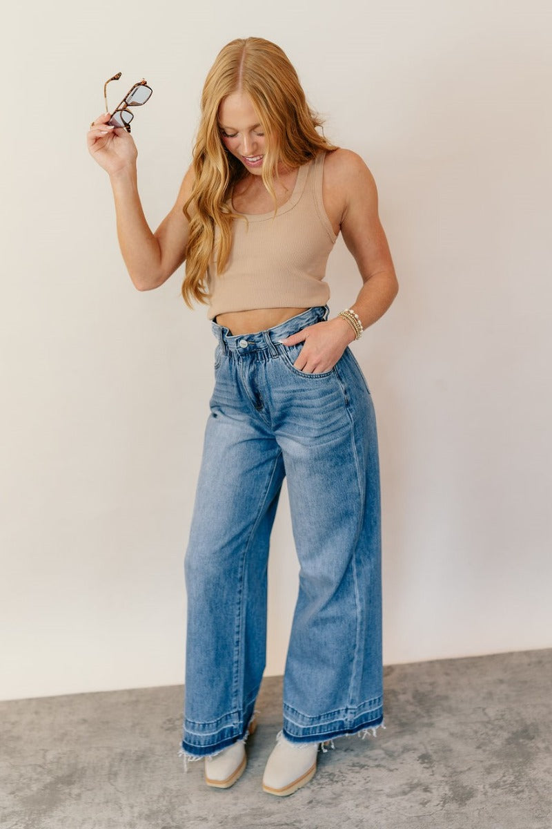 Full body front view of model wearing the Ceros: Billie Wide Leg Paper Bag Jeans that have medium wash denim fabric, pockets, a front zipper, belt loops, an elastic paperbag waist, and wide legs with raw hems.