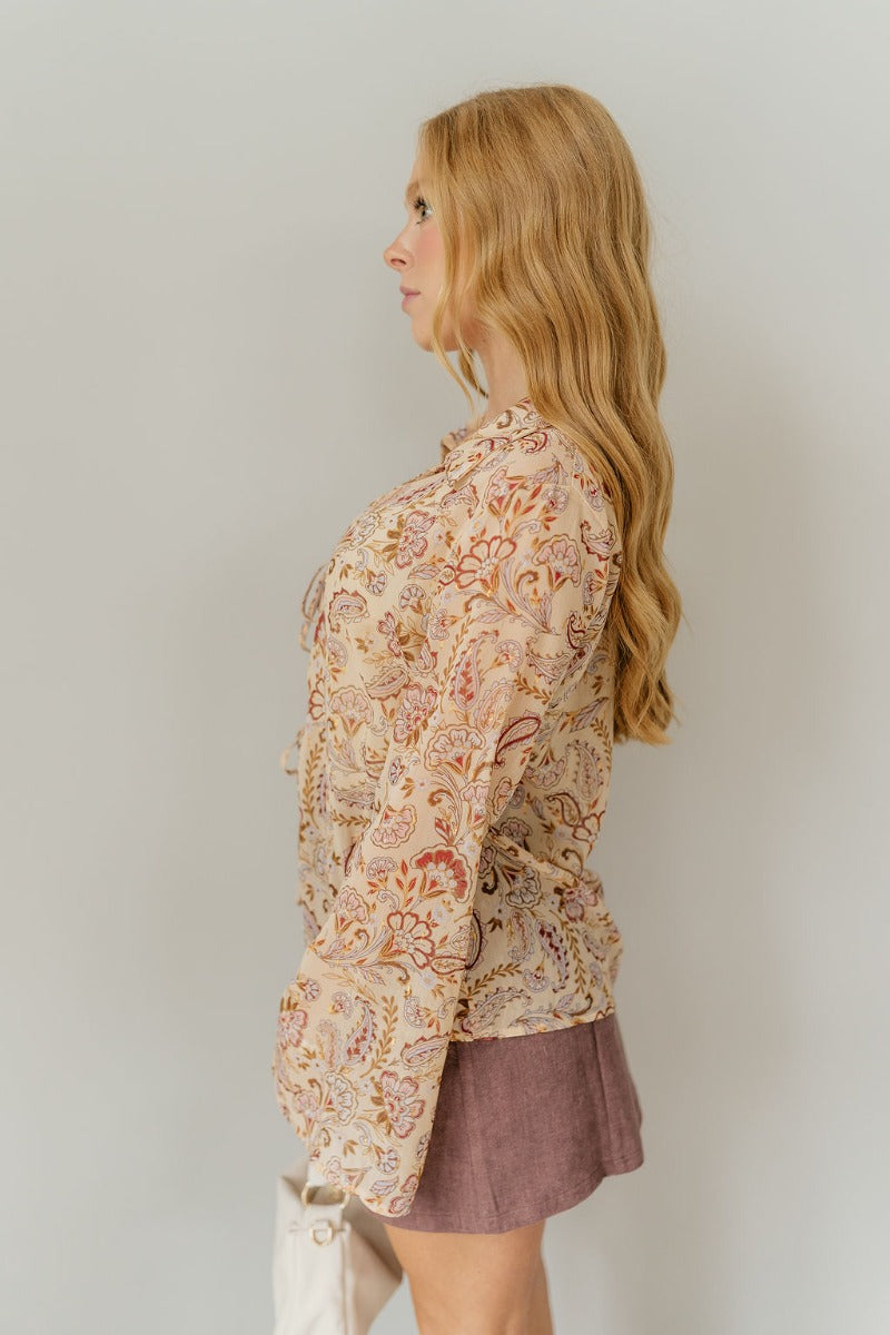 Side view of model wearing the Rosalee Printed Tie-Front Long Sleeve Top that has sheer, a paisley and flroal pattern with gold and silver metallic details, front tie closures, a collared neck, and long flare sleeves with slits.