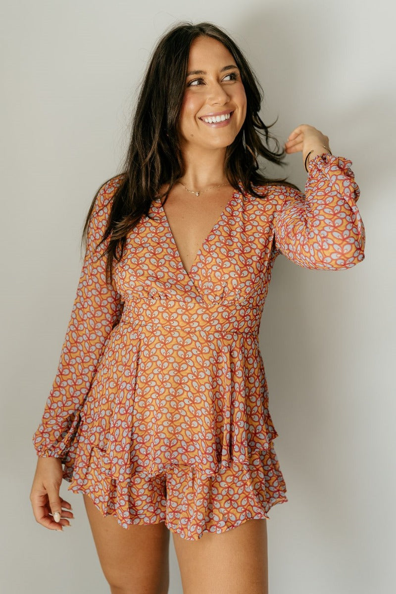 front view of model wearing the Jane Mustard Printed Long Sleeve Romper that has sheer fabric with a mustard, red, light blue and cream pattern, a ruffle tiered design, a plunge neck, and long sleeves.