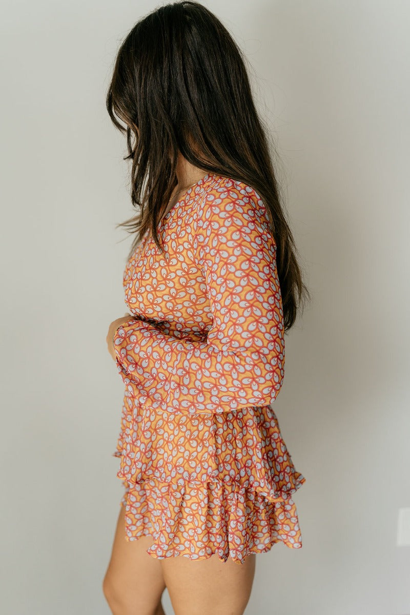 Side view of model wearing the Jane Mustard Printed Long Sleeve Romper that has sheer fabric with a mustard, red, light blue and cream pattern, a ruffle tiered design, a plunge neck, and long sleeves.