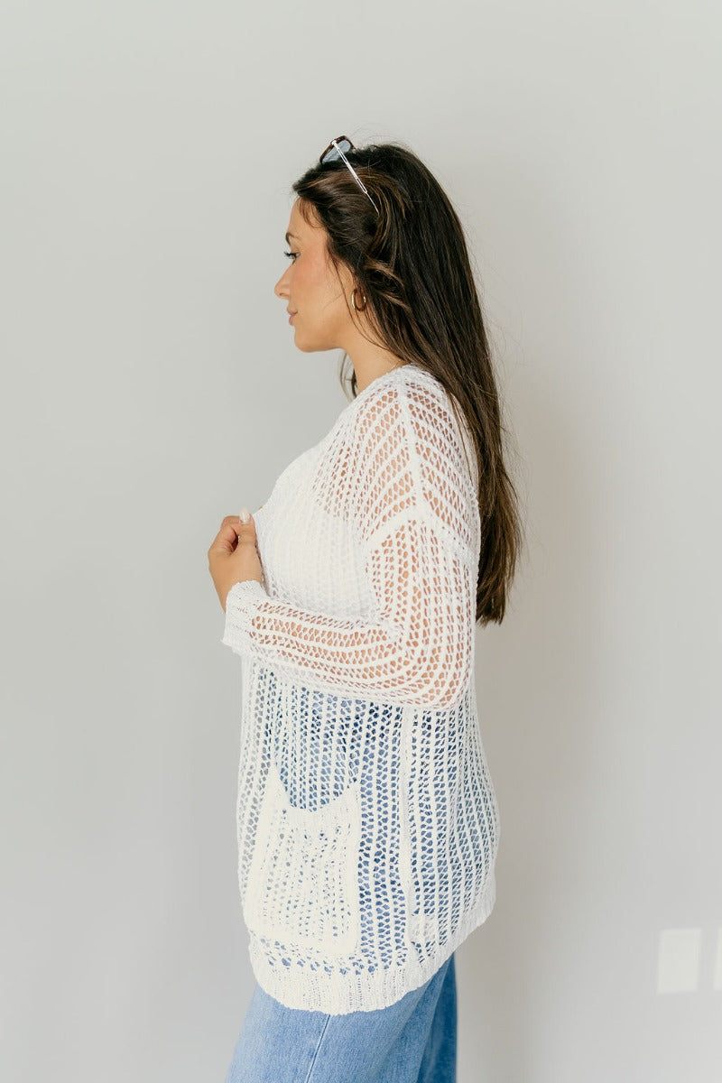 Side view of model wearing the Remington Ivory Crochet Knit Cardigan that has ivory open knit fabric, a front opening with no closure, a thick hem, two front pockets, and long sleeves.