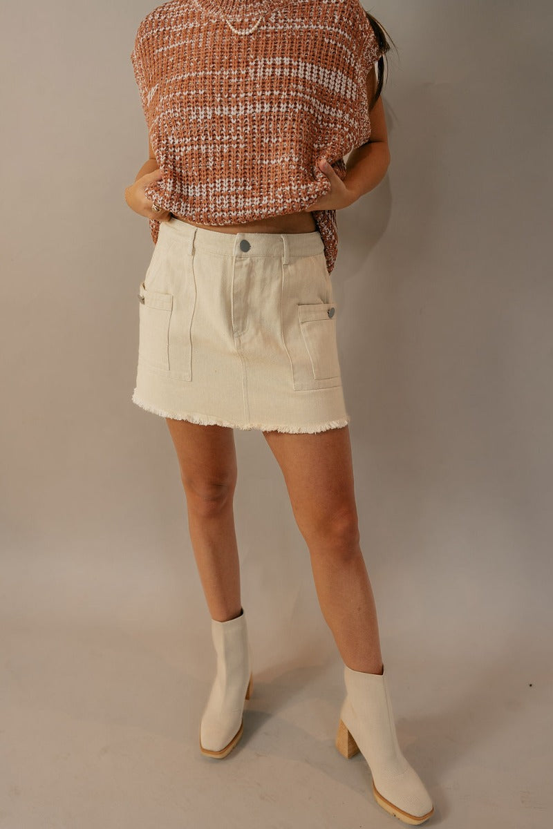 Front view of model wearing the Stella Cream Denim Fray Hem Mini Skir which features ivory denim fabric, a front zipper with a button closure, belt loops, two front pockets, two cargo pockets with faux buttons, and frayed hems.