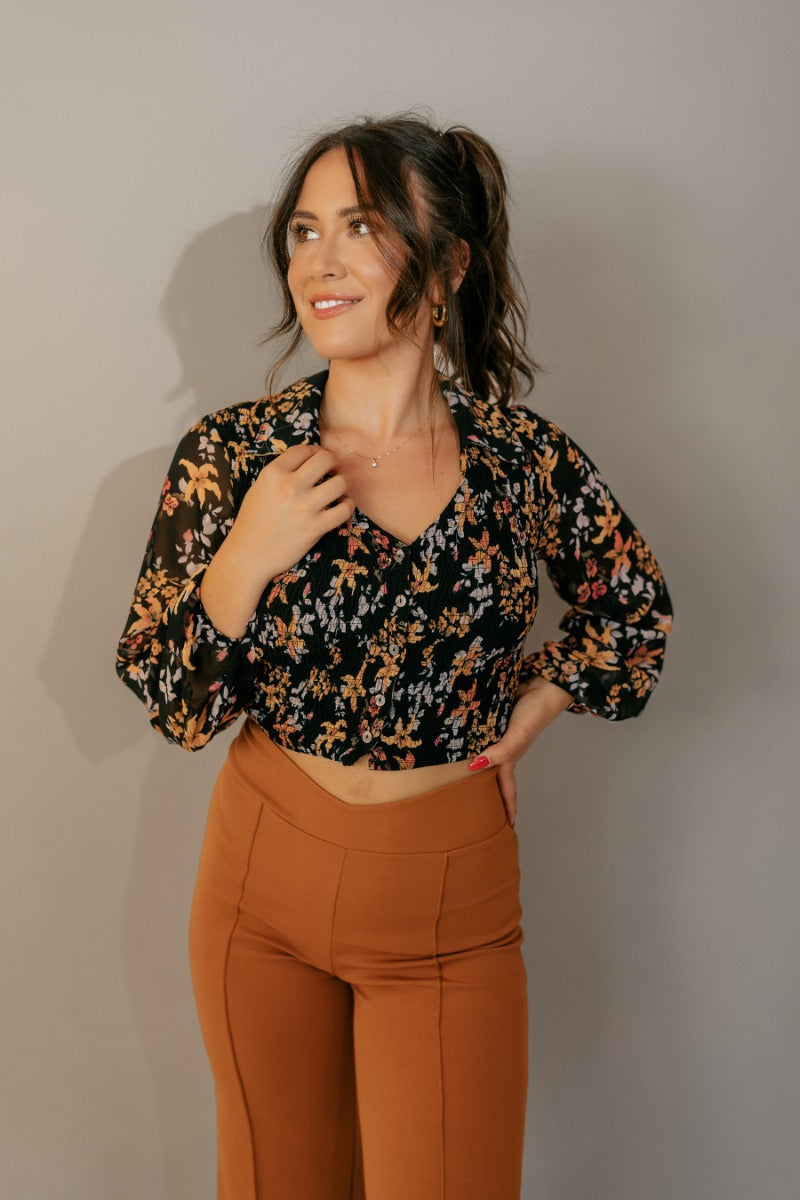 front view of model wearing the Cecilia Black Floral Long Sleeve Cropped Top that has black sheer fabric with a rfloral print, a smocked body, cropped waist, black buttons, a plunge neck, and long balloon sleeves.