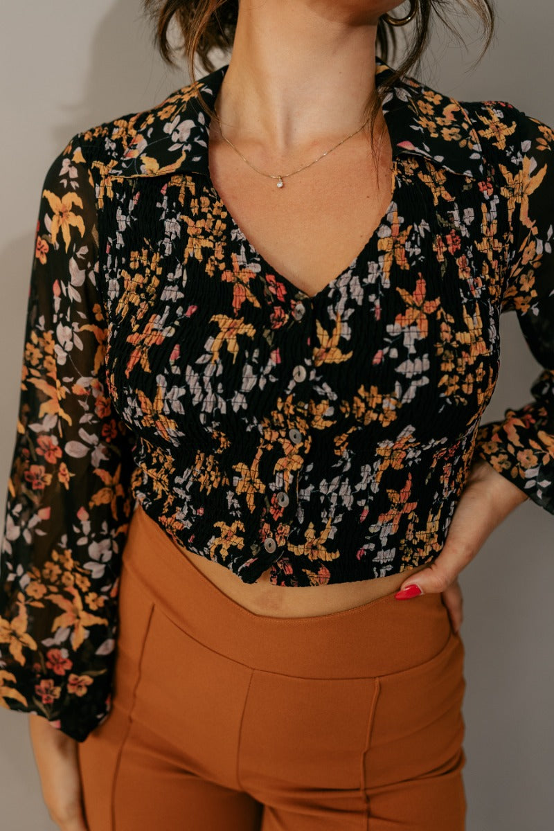 Close front view of model wearing the Cecilia Black Floral Long Sleeve Cropped Top that has black sheer fabric with a rfloral print, a smocked body, cropped waist, black buttons, a plunge neck, and long balloon sleeves.