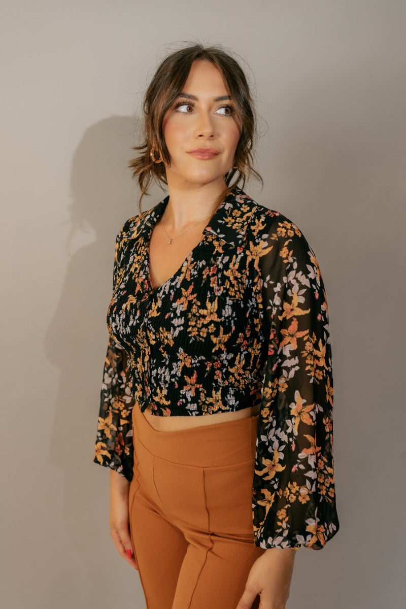 Side view of model wearing the Cecilia Black Floral Long Sleeve Cropped Top that has black sheer fabric with a rfloral print, a smocked body, cropped waist, black buttons, a plunge neck, and long balloon sleeves.