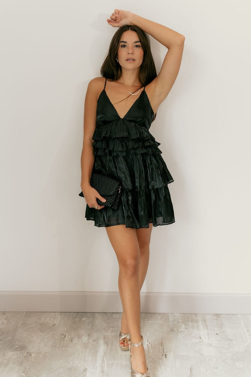 Full body view of model wearing the Everly Black Ruffle Tiered Sleeveless Mini Dress which features black sheer fabric, two tiered style, ruffle details, black lining, a plunge neckline, adjustable straps, and an open back with a tie closure.
