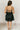 Full body back view of model wearing the Everly Black Ruffle Tiered Sleeveless Mini Dress which features black sheer fabric, two tiered style, ruffle details, black lining, a plunge neckline, adjustable straps, and an open back with a tie closure.