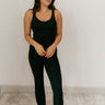 Full body front view of model wearing the Yasmin Black Athletic Onesie Jumpsuit that has black athleisure fabric, monochrome stitched details, a scooped neckline, thick straps, and flared pant legs.