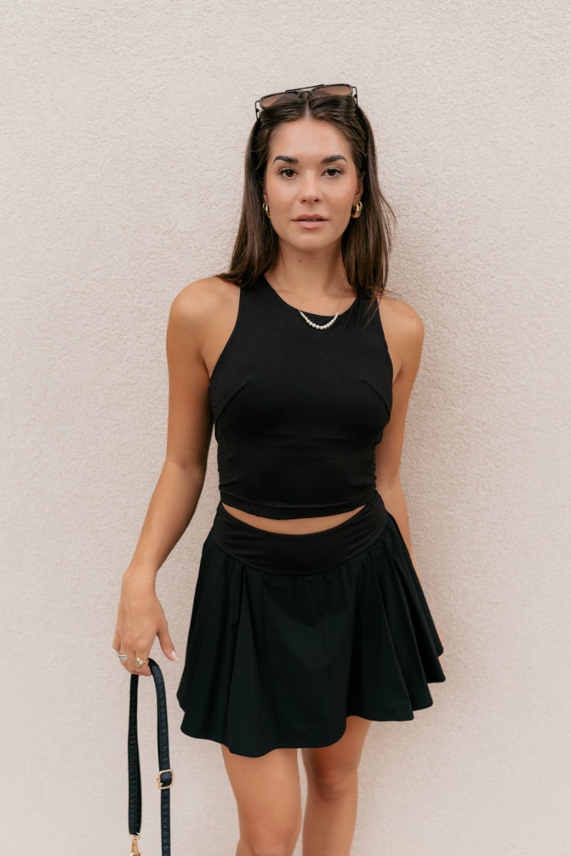 Front view of model wearing the Serena Black Pleated Athletic Skort that has black athleisure fabric, pleated details, black shorts lining with pockets, and a thick waistband.