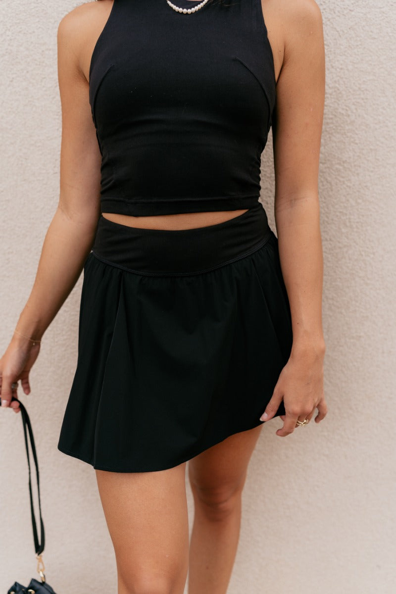 front view of model wearing the Serena Black Pleated Athletic Skort that has black athleisure fabric, pleated details, black shorts lining with pockets, and a thick waistband.