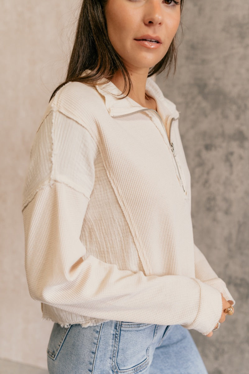 Close side view of model wearing the Brylee Cream Mixed Fabric Half-Zip Top that has cream waffle knit and gauze fabric, textured thread details, a half zip-up neck, dropped shoulders, and long sleeves.