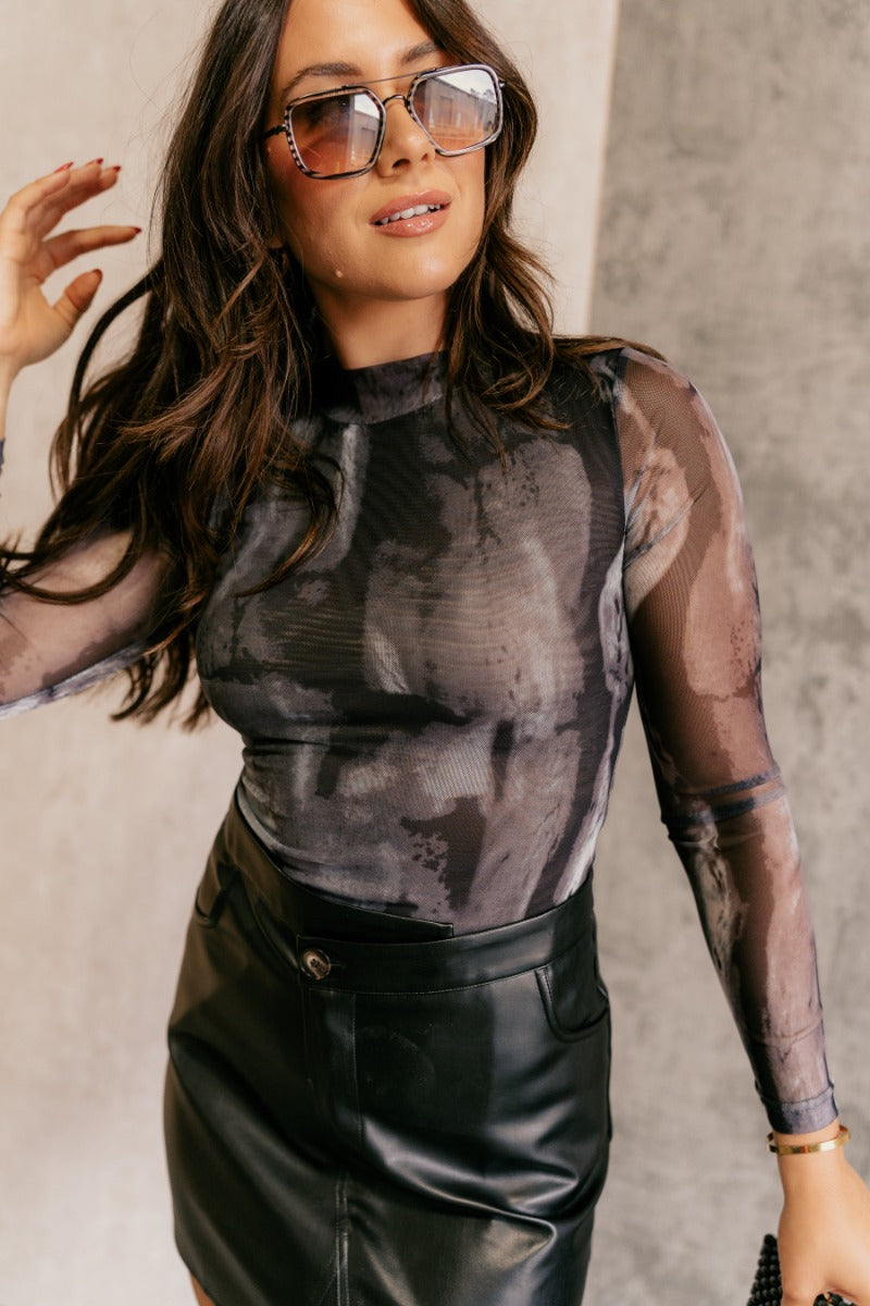front view of model wearing the Leanna Black Marbled Mesh Long Sleeve Bodysuit that has black, grey and white mesh fabric with a watercolor design, a high neck, long sleeves, and a thong bottom.