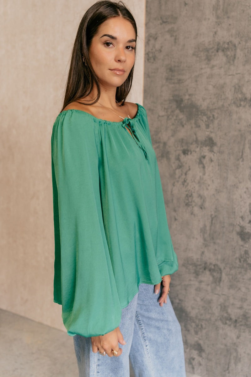Side view of model wearing the Audrey Green Satin Adjustable Neckline Long Sleeve Top which features green satin fabric, a round neckline with a drawstring tie, and long balloon sleeves with elastic wrists.