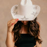 Front view of model wearing the Eloise Ivory & Rhinestone Cowgirl Hat that has a cowboy shape, ivory suede fabric, a rhinestone design on the bottom of hat, and rhinestone star designs on top of the hat.