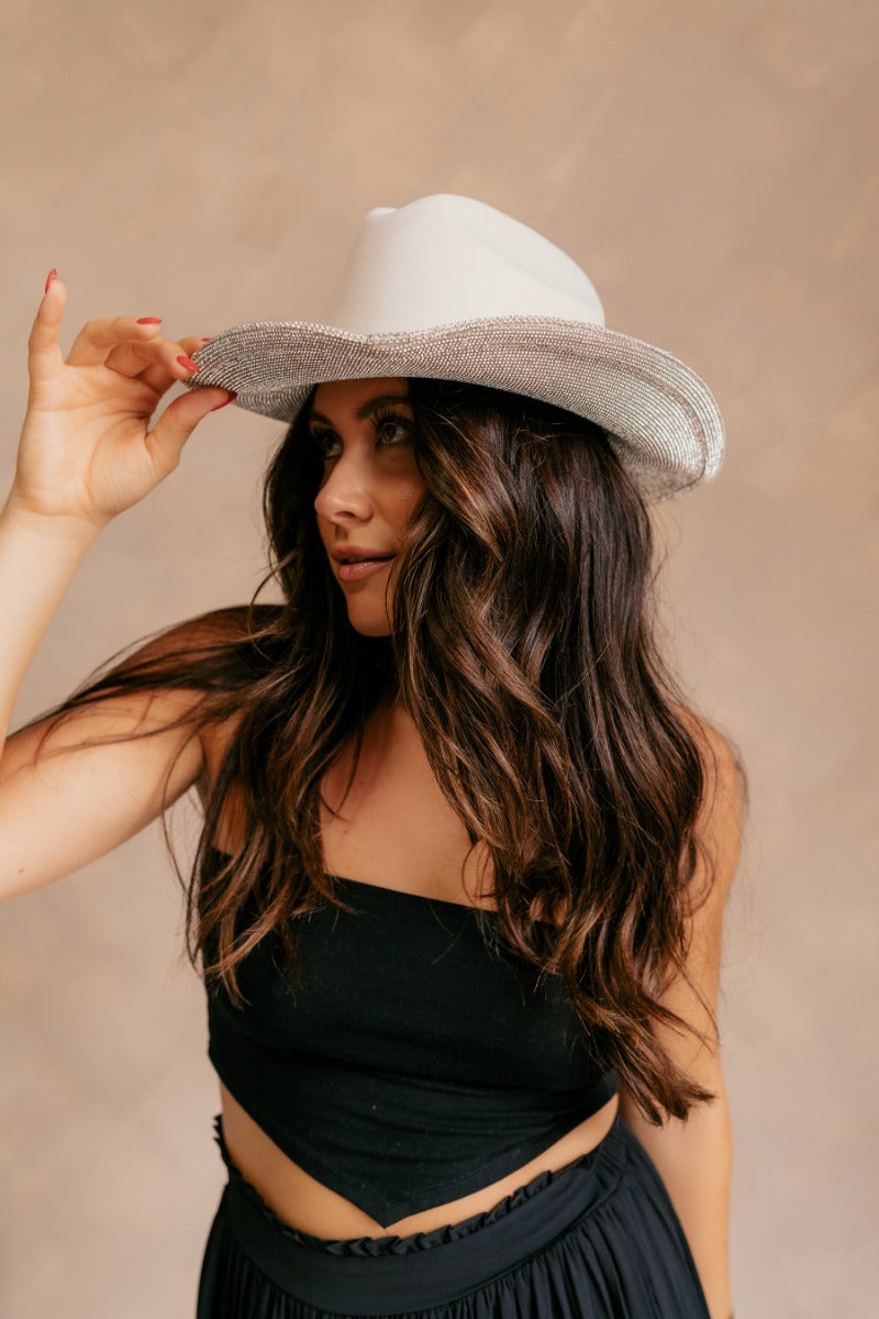 Side view of model wearing the Eloise Ivory & Rhinestone Cowgirl Hat that has a cowboy shape, ivory suede fabric, a rhinestone design on the bottom of hat, and rhinestone star designs on top of the hat.