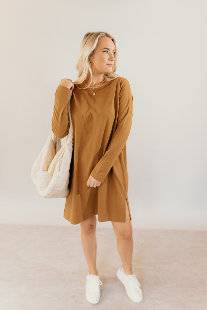 Full body front view of model wearing the Sara Mustard Long Sleeve T-Shirt Dress that has light brown cotton fabric, mini length, slits on each side, a round neckline, dropped shoulders, and long sleeves.