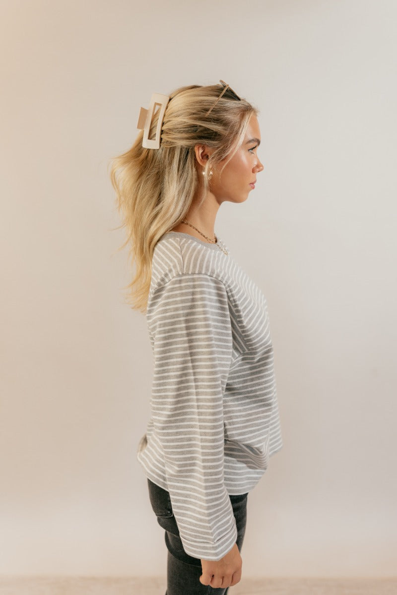 Side view of model wearing the Samantha Grey & White Striped Long Sleeve Top that has grey and white knit fabric, stripe pattern, slits, a round neck, and long flare sleeves.