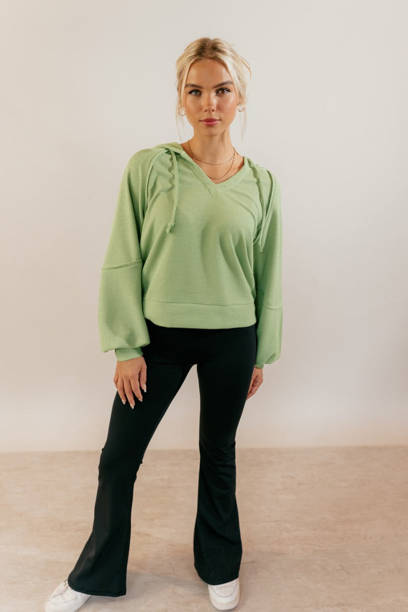 Full body front view of model wearing the Leila Sage Green Hoodie Sweater that has sage textured fabric, a thick hem, a v-neckline with a hood and drawstring ties, and long puff sleeves with cuffs.