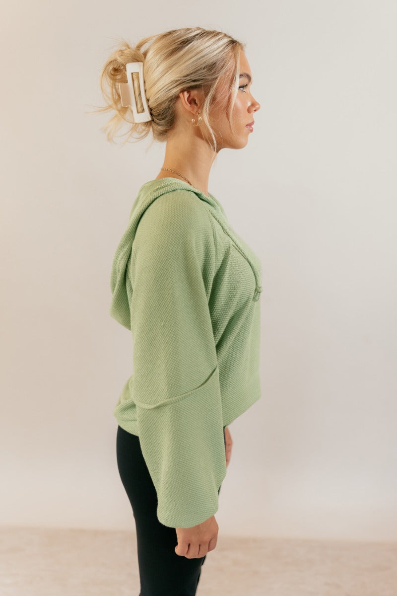 Side view of model wearing the Leila Sage Green Hoodie Sweater that has sage textured fabric, a thick hem, a v-neckline with a hood and drawstring ties, and long puff sleeves with cuffs.