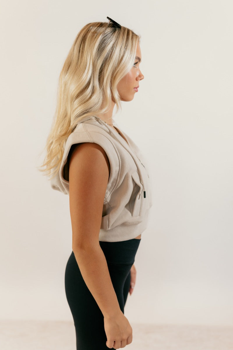 Side view of model wearing the Melanie Taupe Sleeveless Cropped Hoodie that has taupe knit fabric, a cropped waist, pockets, a front zipper a hood, and a sleeveless design.