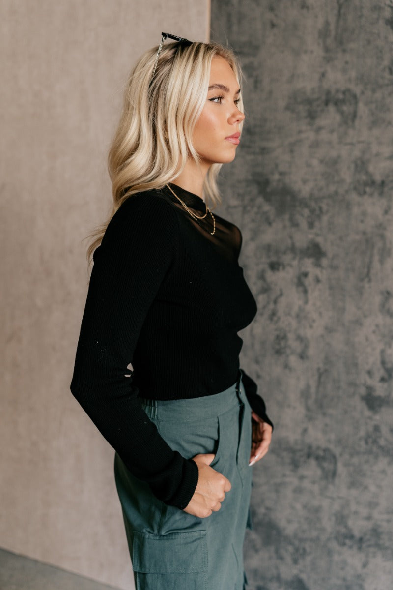 Side view of model wearing the Callie Black Mesh Detail Long Sleeve Top that features black ribbed knit and black mesh fabric, a high neckline, and long sleeves.