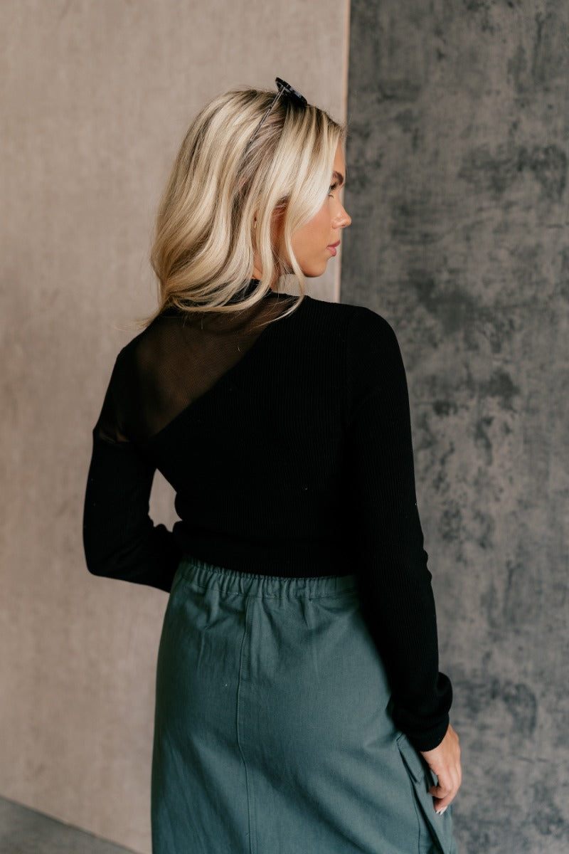 Back view of model wearing the Callie Black Mesh Detail Long Sleeve Top that features black ribbed knit and black mesh fabric, a high neckline, and long sleeves.
