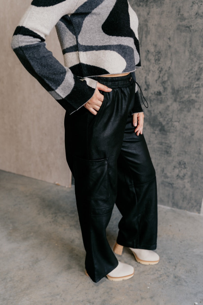 Side view of model wearing the Zuri Black Faux-Leather Pants that have black soft faux leather fabric, two front pockets, an elastic waistband with a drawstring tie, and wide legs.