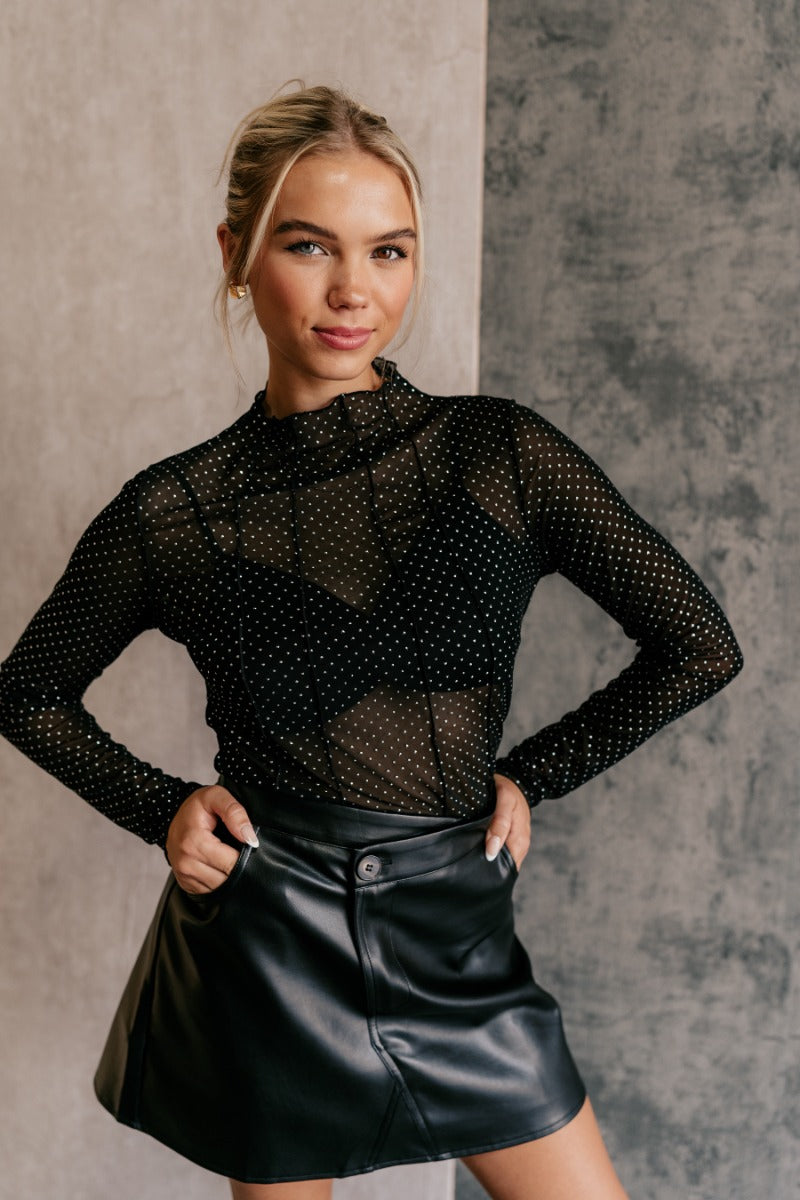 front view of model wearing the Anastasia Black Embellished Mesh Top that has black mesh fabric, silver embellishments, exposed seam details, a high neckline, and long sleeves.