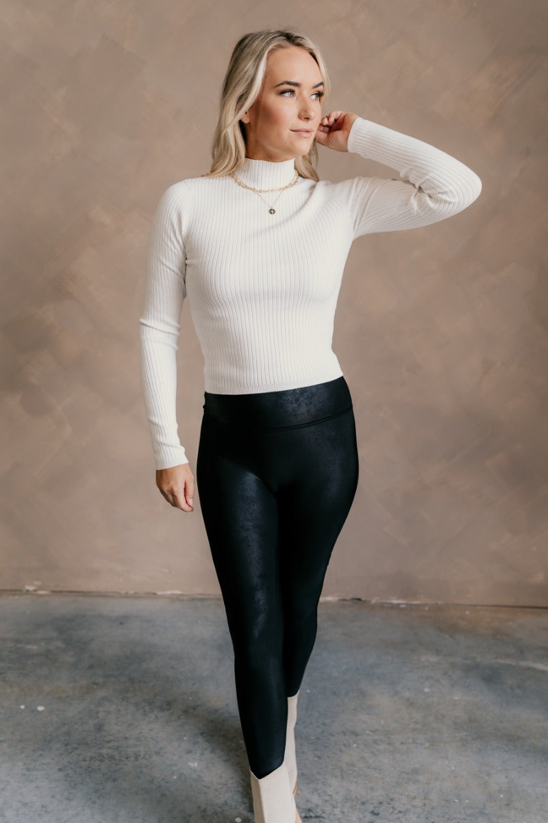 Full body front view of model wearing the Norah Off White Mock Neck Top that has off white ribbed fabric, a high neckline, and long sleeves.