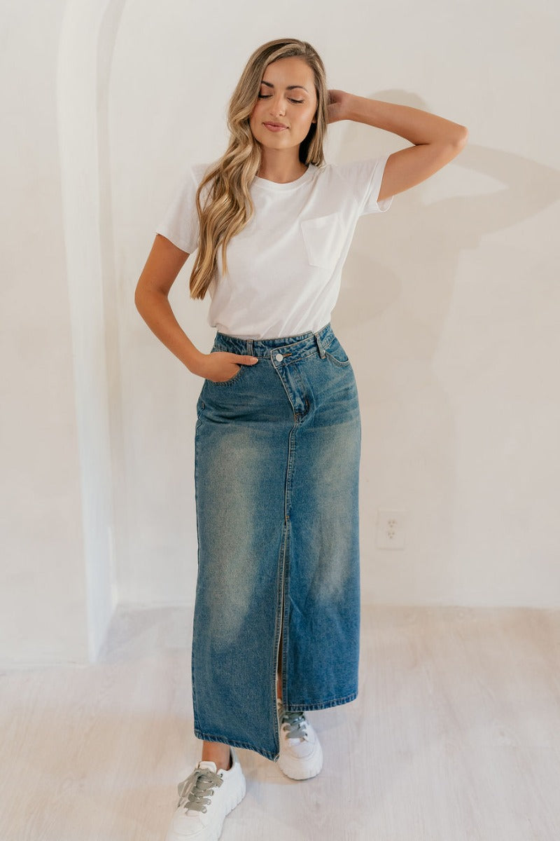 Full body view of model wearing the Skye Blue Denim Slit Midi Skirt which features washed blue denim fabric, a front slit, a front slanted zipper with a button closure, belt loops, front pockets, back pockets, and maxi length.