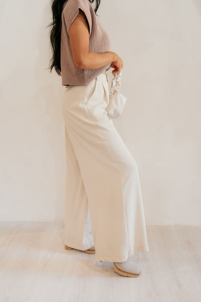 Side view of model wearing the Scarlett Beige Wide Leg Pants that have light beige lightweight fabric, two front pockets, a front zipper with a hook closure, belt loops, and wide legs.