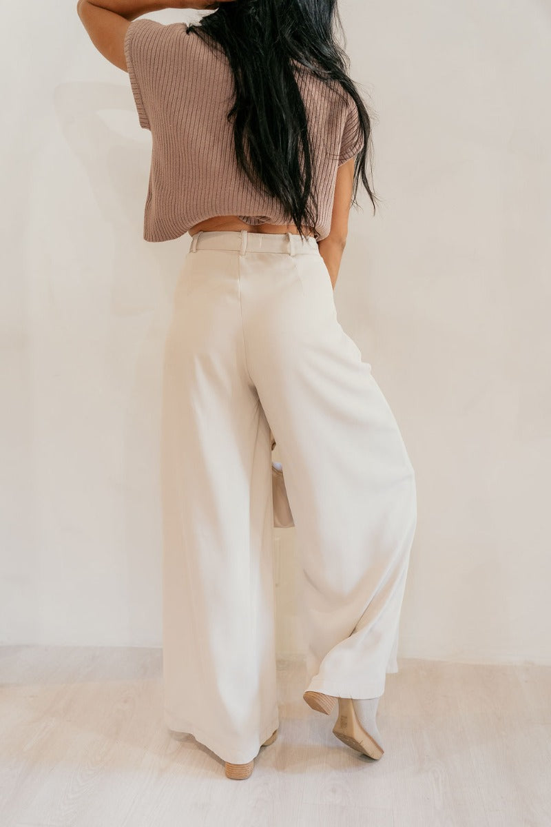 Back view of model wearing the Scarlett Beige Wide Leg Pants that have light beige lightweight fabric, two front pockets, a front zipper with a hook closure, belt loops, and wide legs.