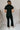 Full body back view of model wearing the Rielle Black Adjustable Belt Short Sleeve Jumpsuit which features black fabric, two front pockets, a front zipper, an adjustable belt with a silver buckle, a collared neckline, short sleeves, and straight pant legs