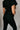 Back view of model wearing the Rielle Black Adjustable Belt Short Sleeve Jumpsuit which features black fabric, two front pockets, a front zipper, an adjustable belt with a silver buckle, a collared neckline, short sleeves, and straight pant legs.