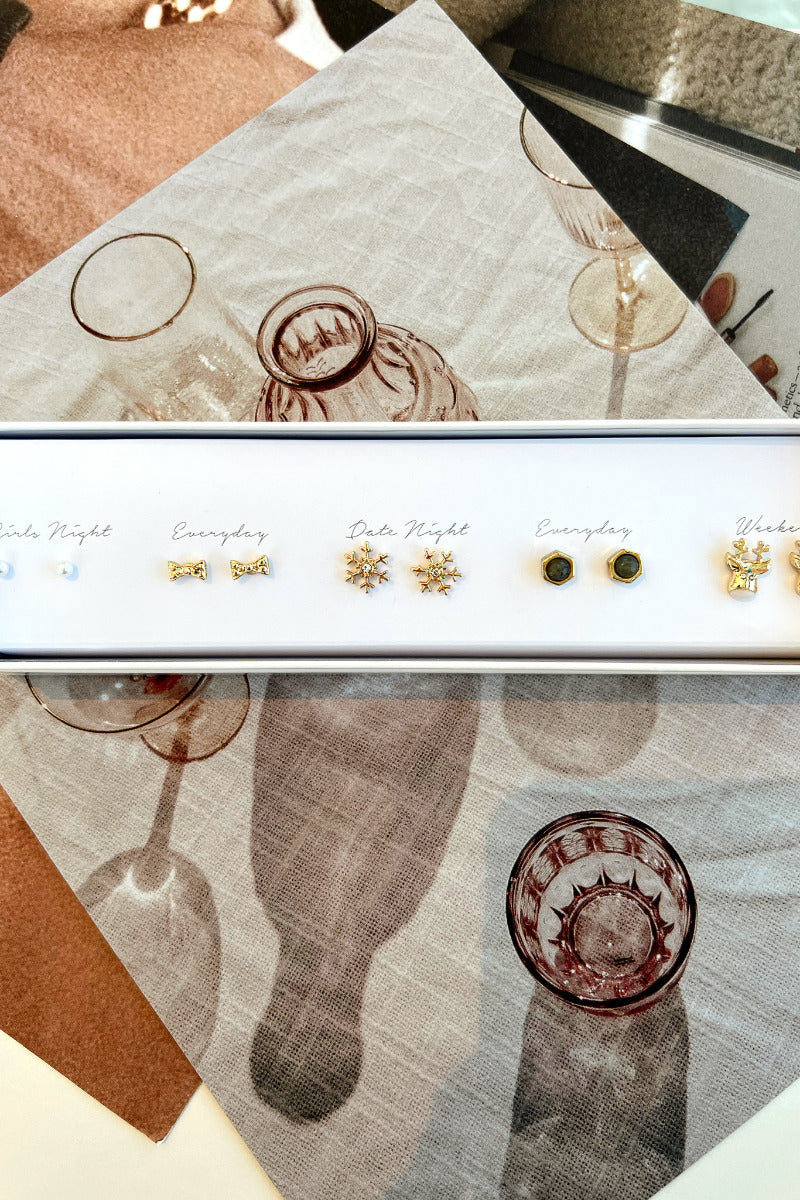 Top view of the Let It Snow Earring Set, packaged in a white box with "Girls night, everyday, date night, everyday, and weekend" text. It includes 5 pairs of stud earrings, featuring pearls, gold bows, gold snowflakes, circle stones, and gold reindeer