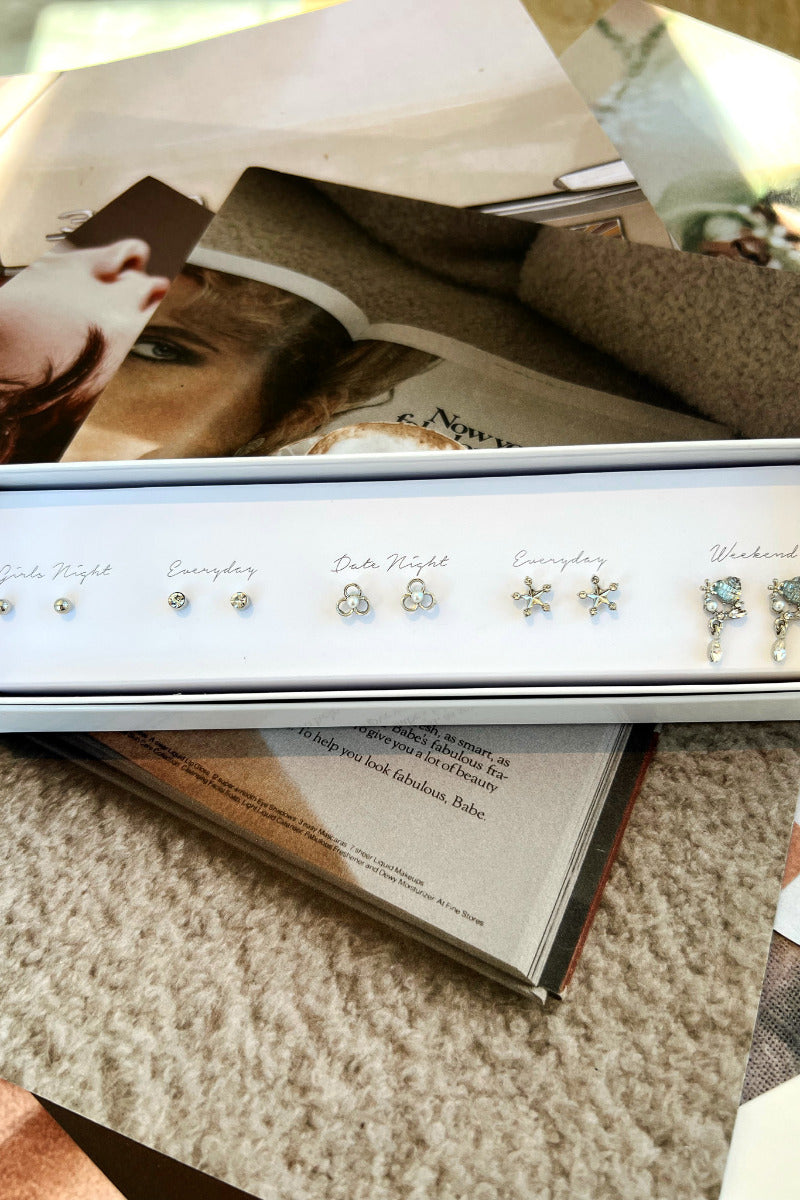Top view of the Snow Day Earring Set, packaged in a white box with "Girls night, everyday, date night, everyday, and weekend" text. It includes 5 pairs of silver stud earrings, features spheres, rhinestones, snowflakes, and tear drops.