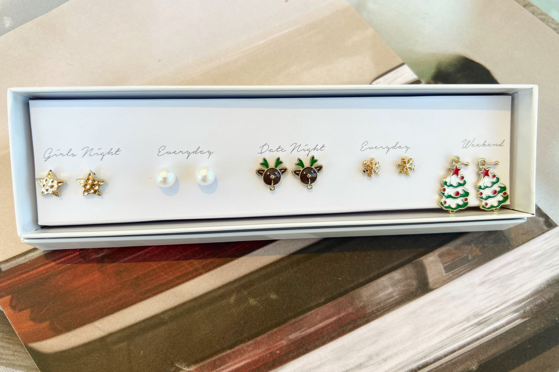 Top view of the Merry and Bright Earring Set, packaged in a white box with "Girls night, everyday, date night, everyday, and weekend" text. It includes 5 pairs of earrings, with gold stars, pearls, reindeer, snowflakes, and Christmas trees