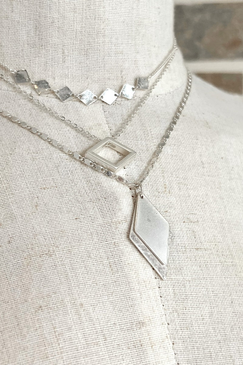 Close up view of the Time To Travel Necklace In Silver which features matte silver triple layer chain link and short length chain with diamond shaped attachments.