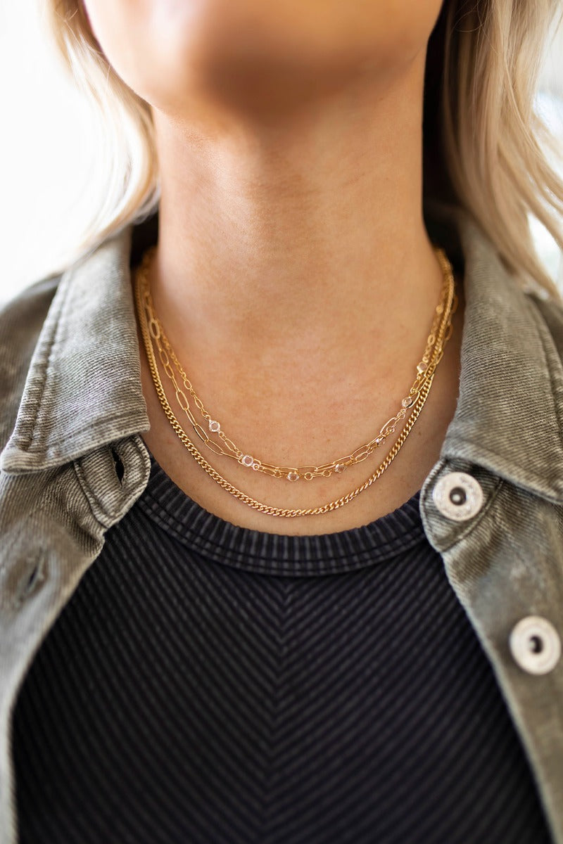 Close up view of model wearing the You're A Gem Necklace which features triple gold chain layer with links and clear stones. 