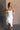 Front view of model wearing the Liliana White Asymmetric Sleeveless Midi Dress that has white knit fabric, an asymetrical ruffle hem, midi length, a square neckline, and adjustable straps.